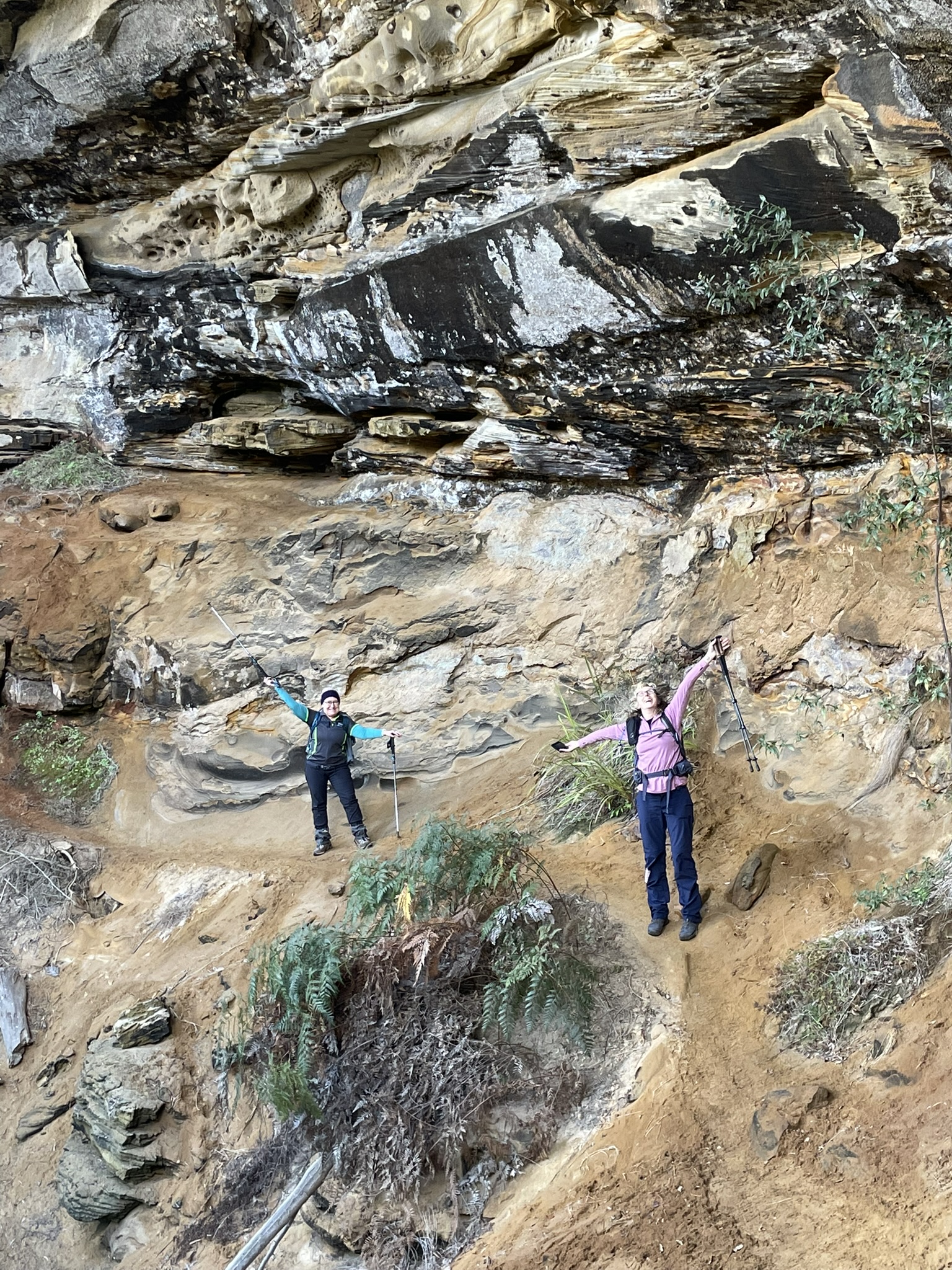 2 hikers with arms stretched to the heavens show us the majestic height of the the sandstone cliff face